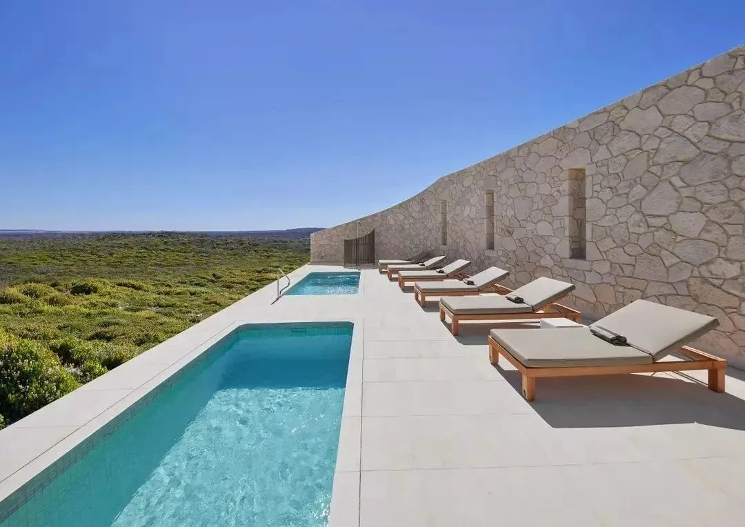 Long rectangular plunge pool beside a stone wall and sun loungers, overlooking coastal bushland