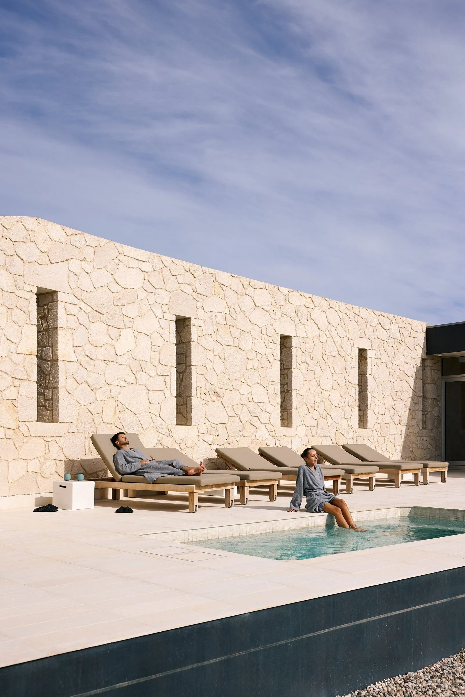 People relaxing on sun loungers beside a rectangular plunge pool and a stone feature wall