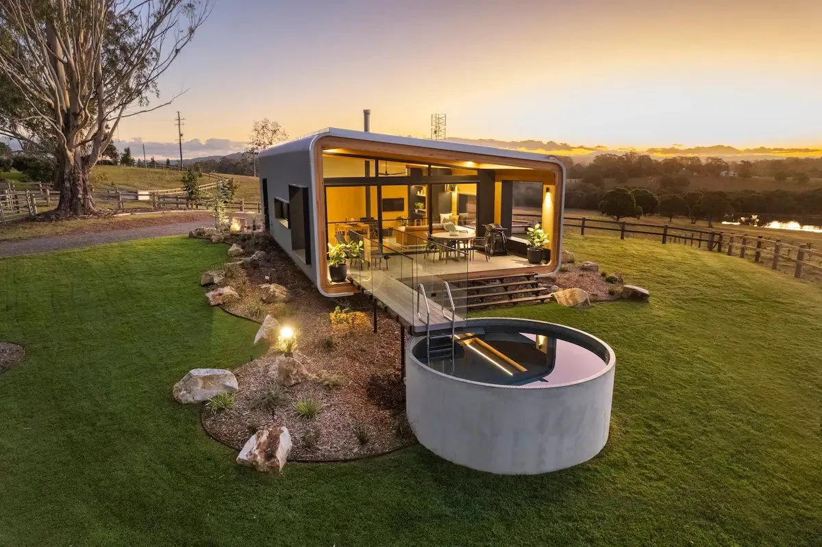 Round plunge pool lit at dusk beside a glass-fronted cabin on a lawn