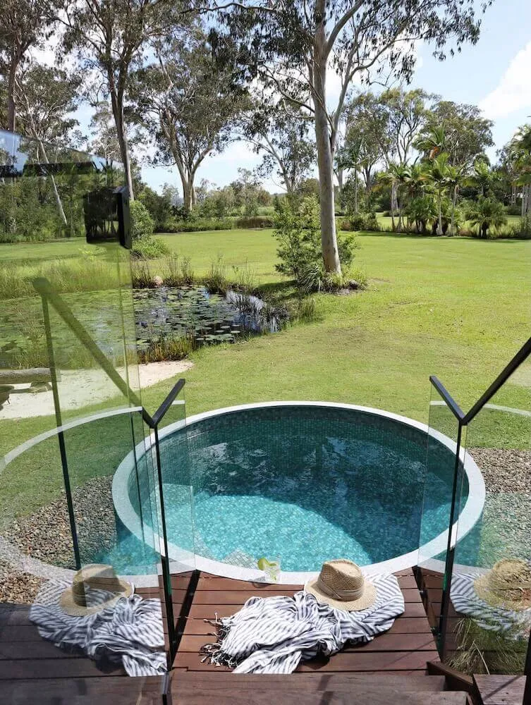 Round plunge pool with glass fencing and a timber step in a large garden