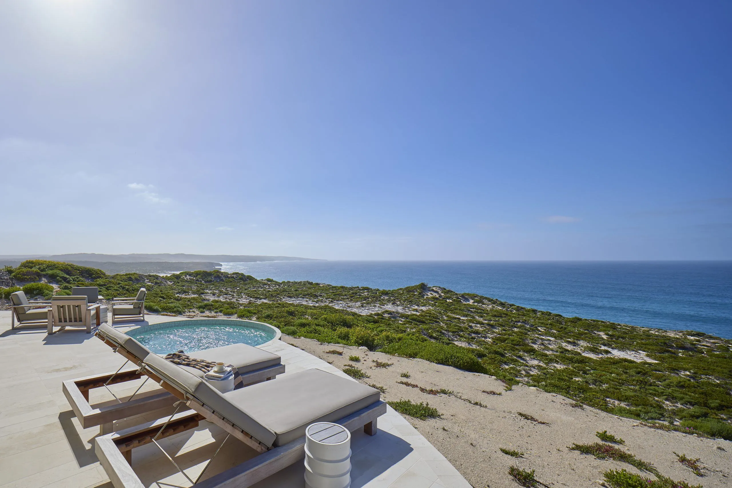 A round plunge pool on a clifftop terrace with sun loungers, overlooking coastal heath and the ocean