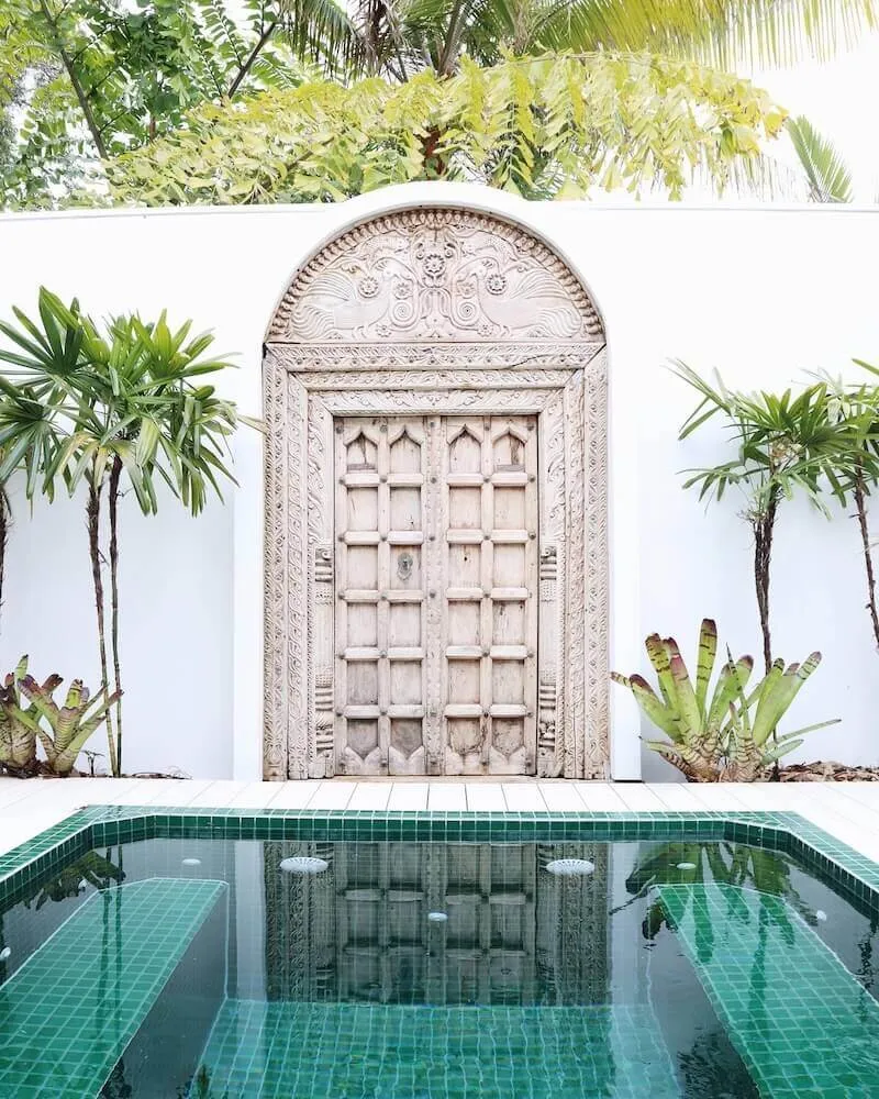 A green-tiled Zen pool in front of a white wall with a carved timber door and palms