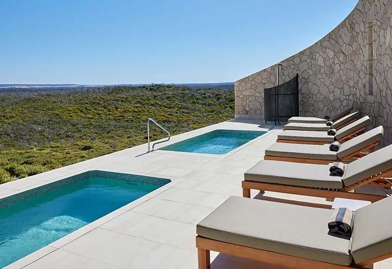 Two rectangular plunge pools beside sun loungers and a curved stone wall, with bushland views