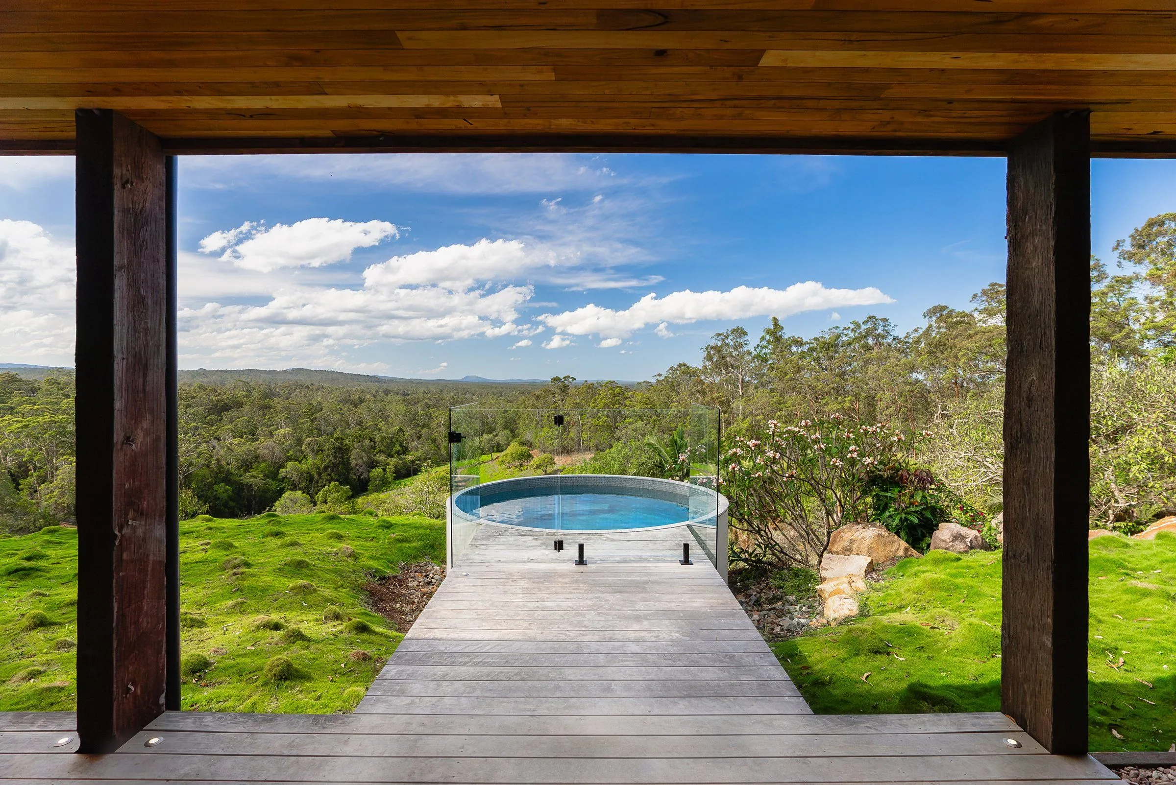 Round plunge pool at the end of a timber boardwalk, framed by a covered deck, with bushland views