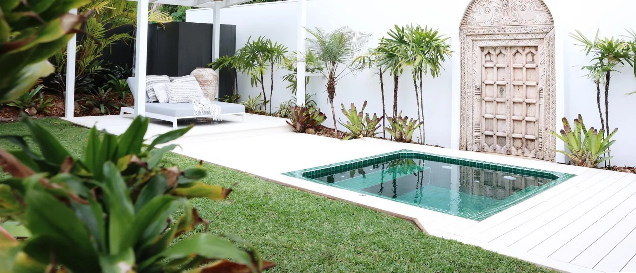 A green-tiled Zen square plunge pool in a white-walled garden with palms and a carved timber door