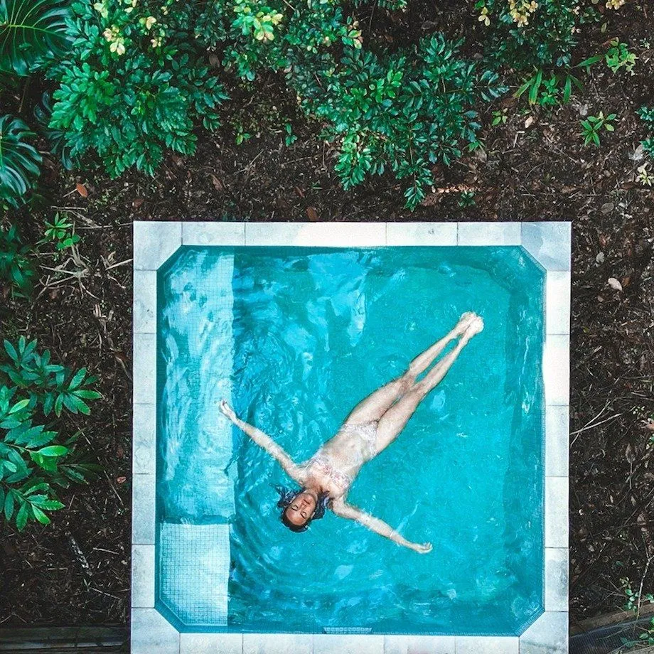 Top-down view of a person floating in a square plunge pool surrounded by greenery