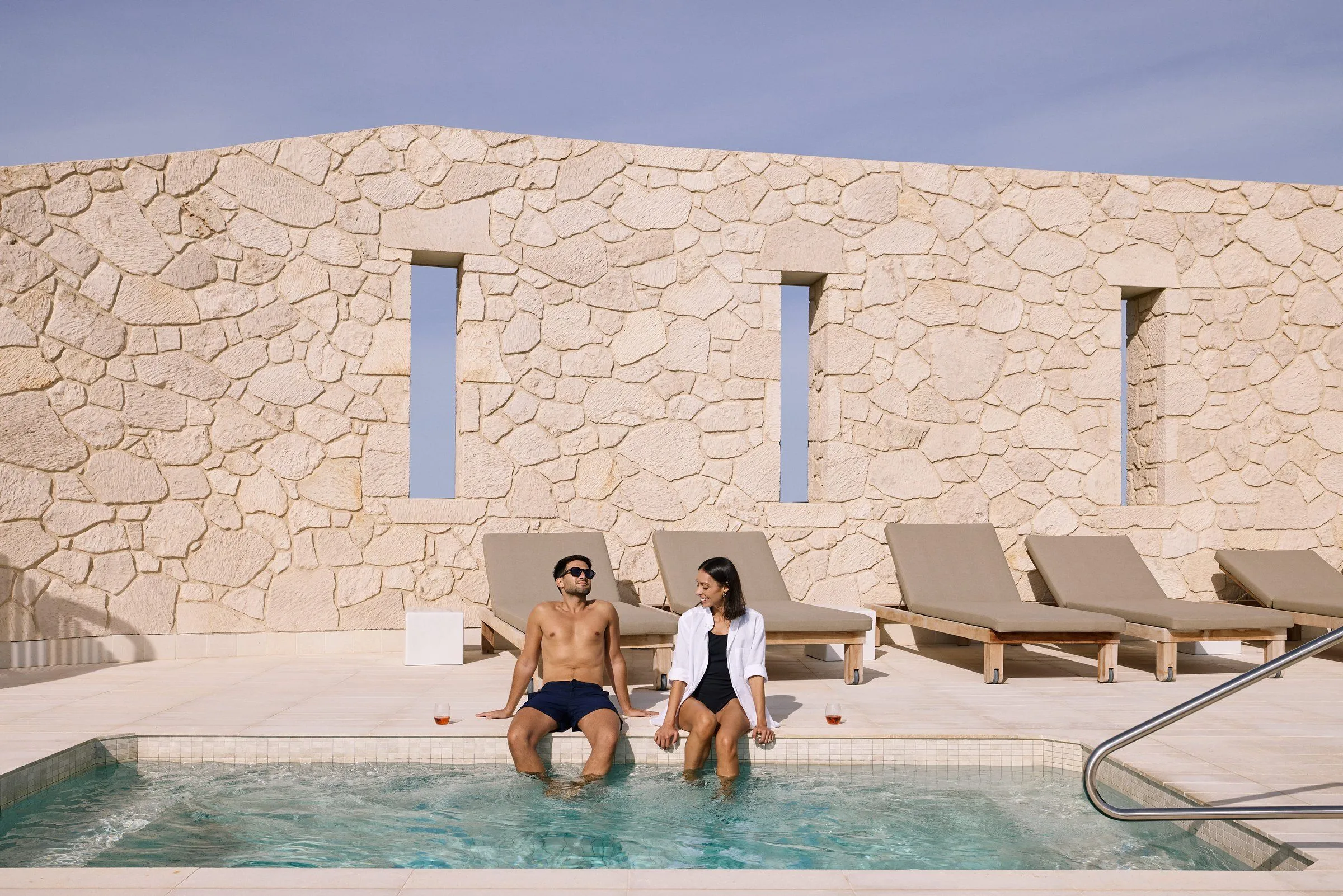 Couple sitting on the edge of a rectangular plunge pool in front of a stone wall