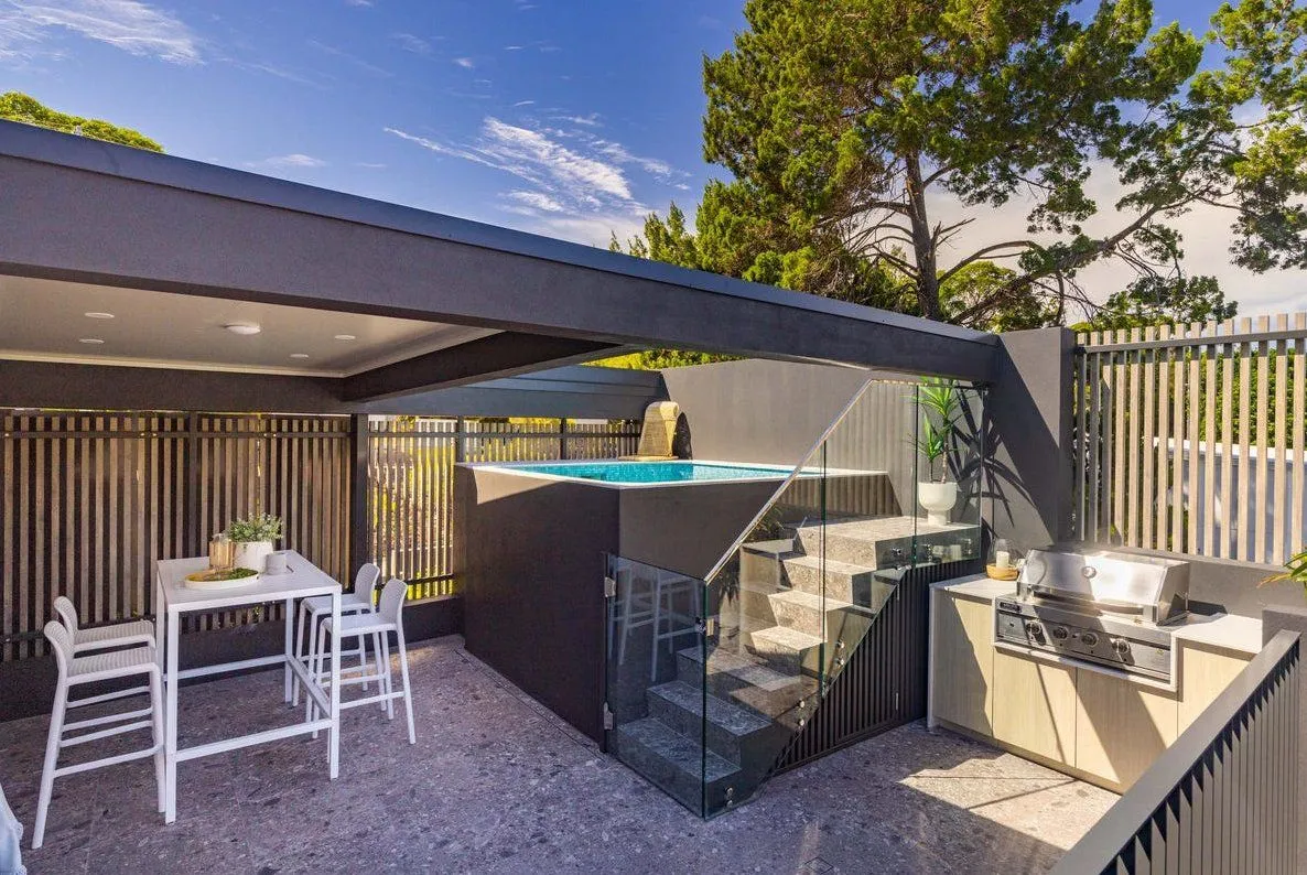 Raised square plunge pool with glass steps beside a covered outdoor kitchen and dining area