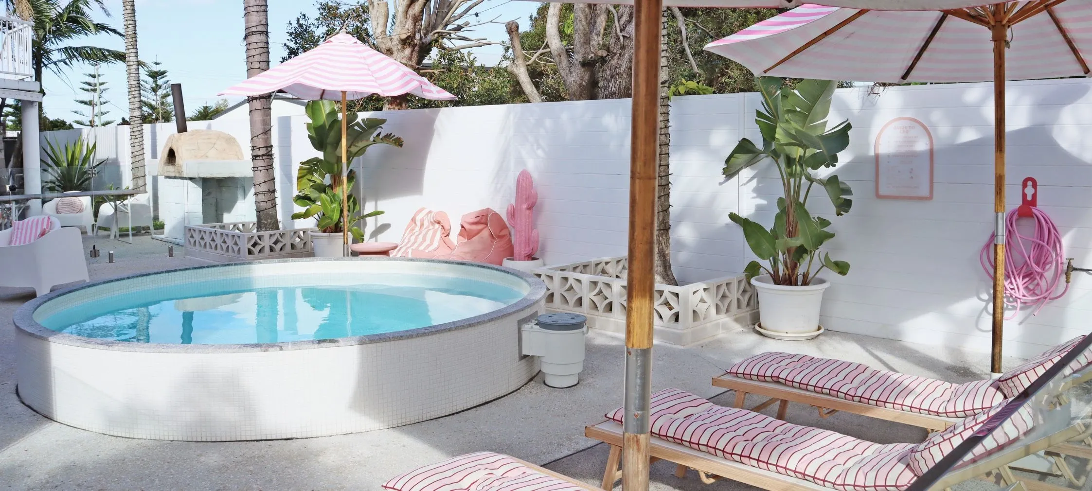 A white round Bliss plunge pool in a courtyard with pink-striped sun loungers and umbrellas